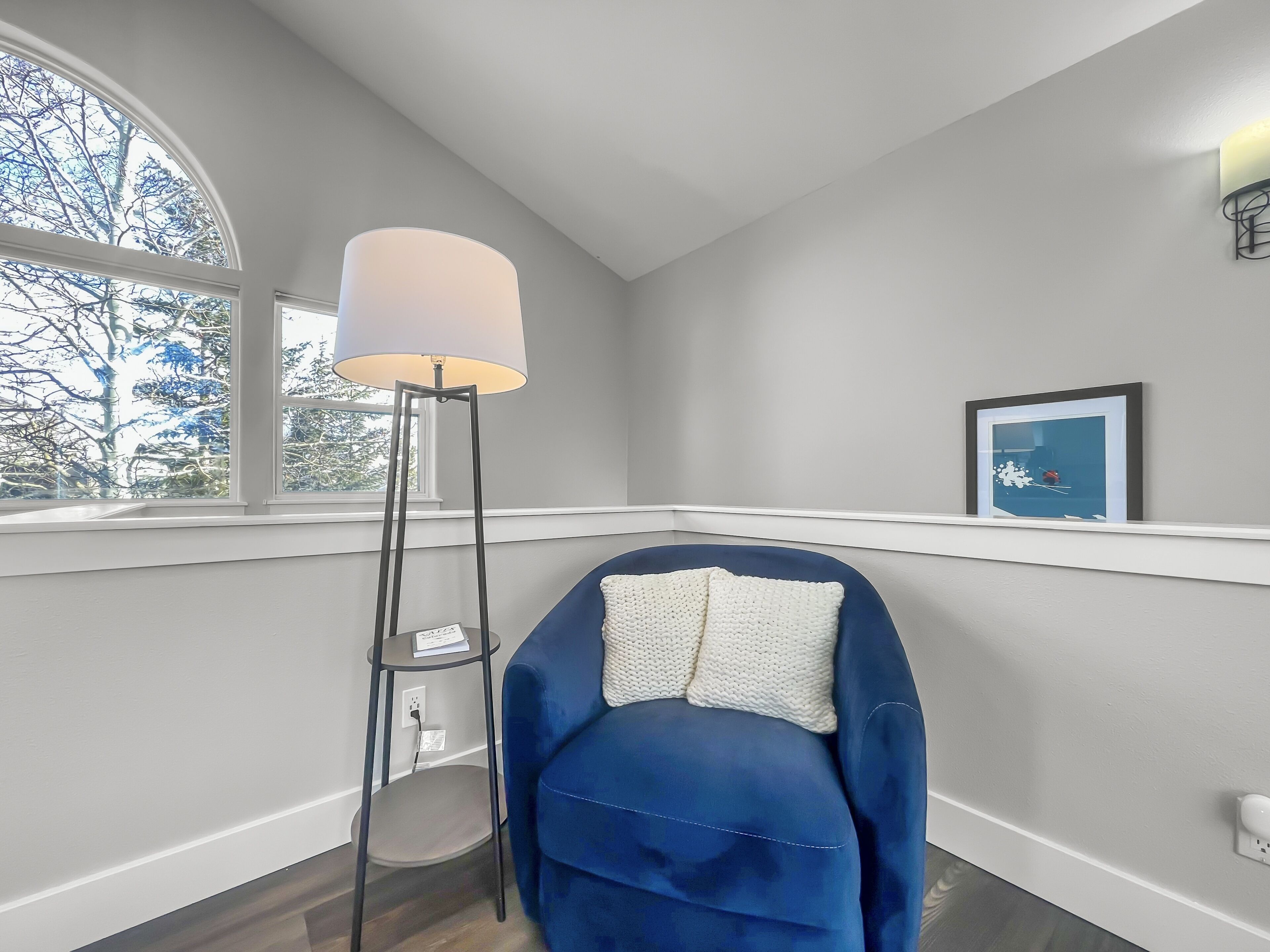 A room with a large window, a floor lamp, a blue chair with a white cushion, and a framed picture on the wall.
