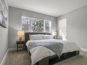 Une chambre à coucher avec un lit, deux lampes de chevet, une fenêtre donnant sur des arbres et un tableau accroché au mur.