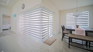 A modern dining area with a chandelier, a dining table with chairs, and a large window with blinds.