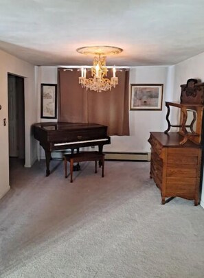 A room with a grand piano, a wooden dresser, a chandelier, and two framed pictures on the wall.