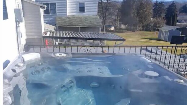 A hot tub on a wooden deck with a metal railing, surrounded by a residential area with houses and trees.