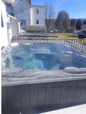 A hot tub on a wooden deck with a metal railing, surrounded by a residential area with houses and trees.