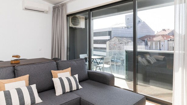 Una sala de estar moderna con un sofá seccional gris, un puerta corredera de vidrio a un balcón y vistas de edificios.