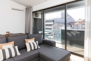 A modern living room with a grey sectional sofa, a glass sliding door to a balcony, and a view of buildings outside.