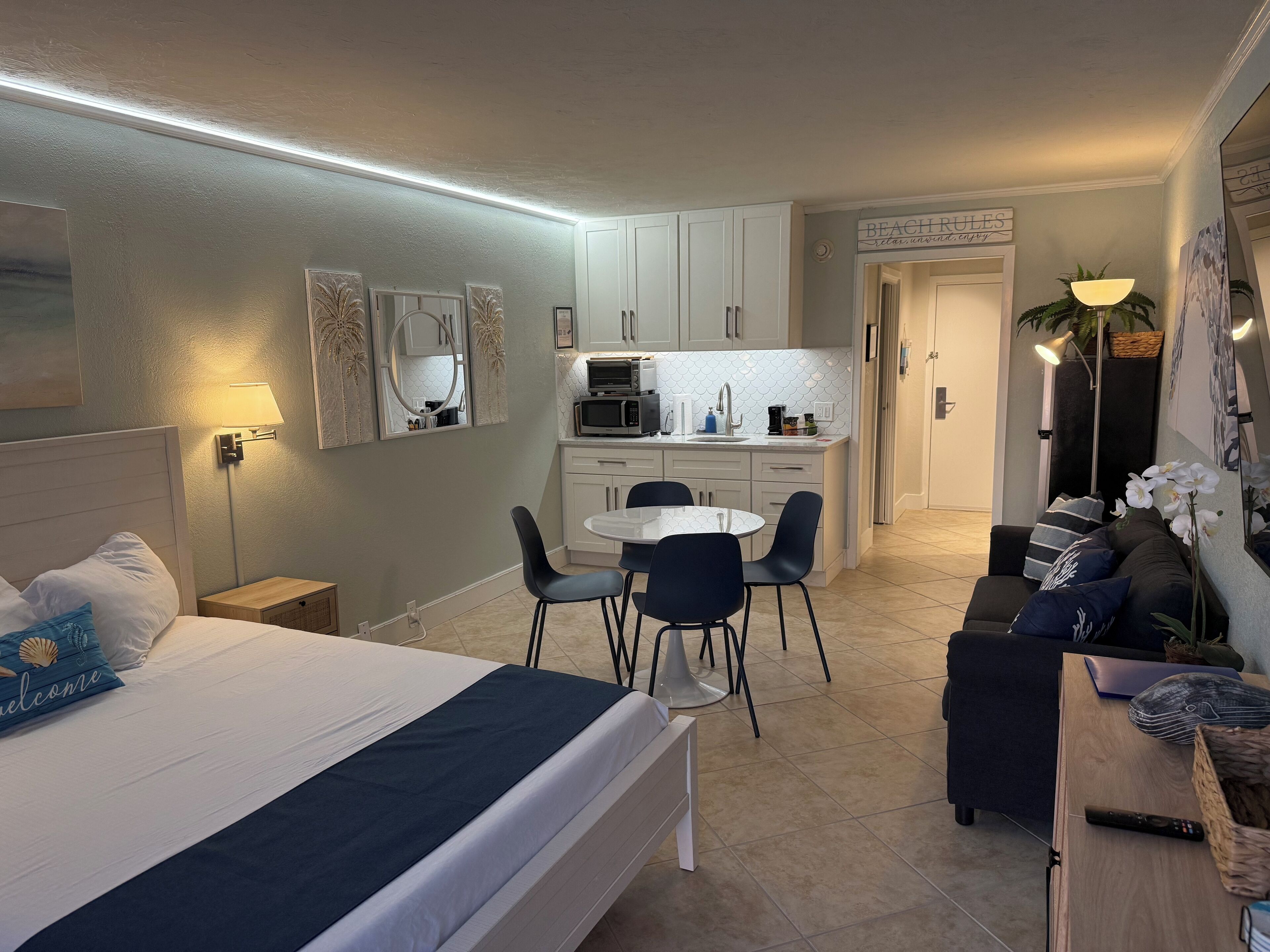 A modern hotel room with a bed, a small desk, a dining table with chairs, a kitchenette, and a sofa.