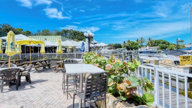An outdoor dining area with tables and chairs, umbrellas, and a view of boats and water. - ALL-NEW Waterfront Room, Heated Pool, Tiki Bar/Grill (Sarasota)