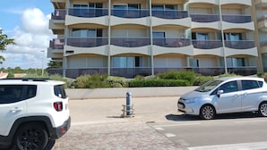 Un edificio de varios pisos con balcones, un espacio pavimentado al frente y dos coches estacionados.