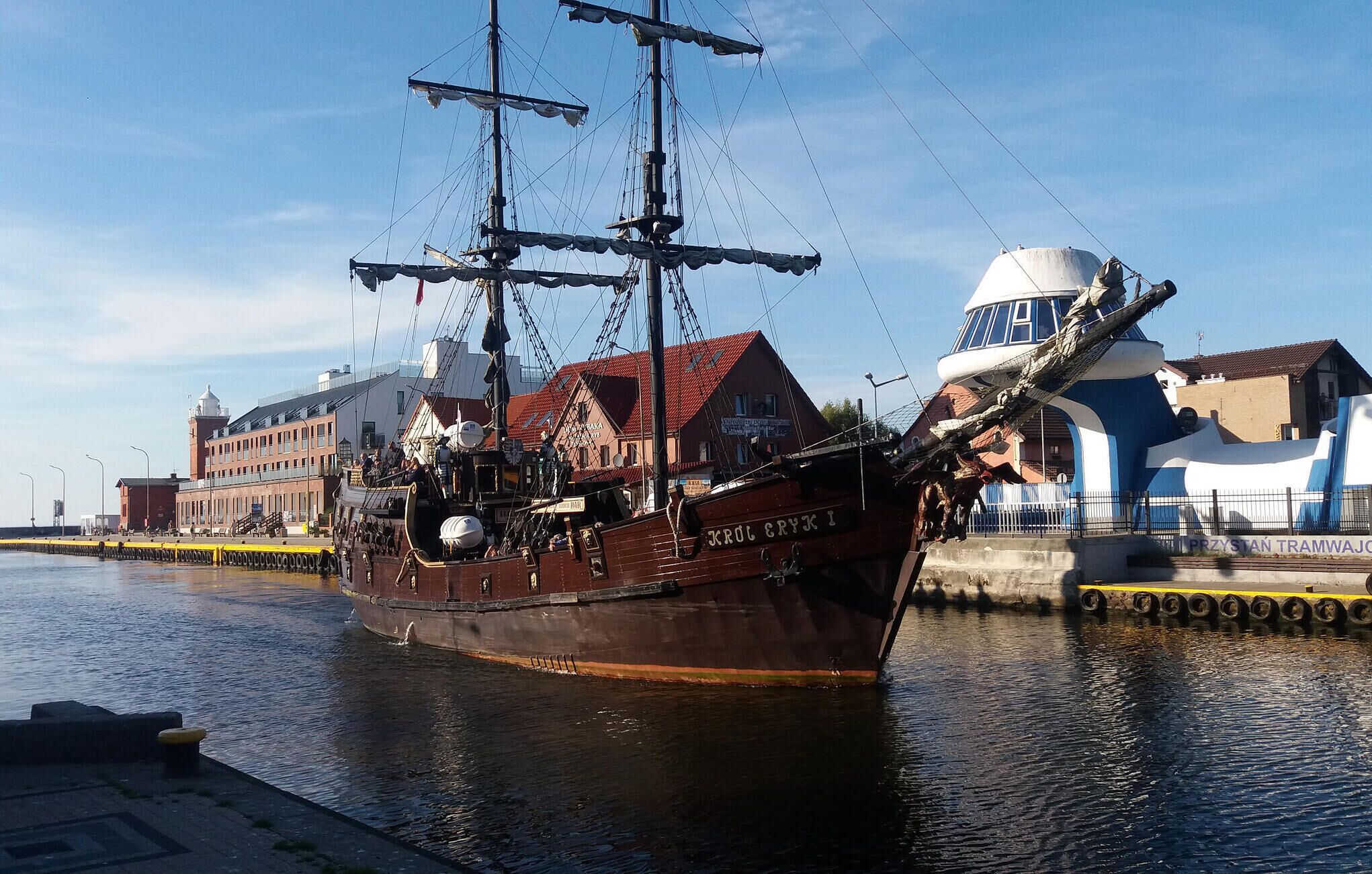 A historic sailing ship docked at a pier with buildings in the background.