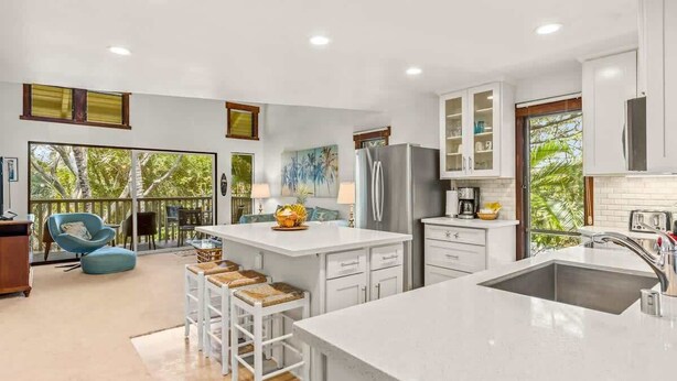 A modern kitchen with white cabinetry, a large island with stools, and a view of the outdoors. - Waikomo Stream Villas 530 – A/C, 2 Bedroom, PBAC Membership (Koloa)