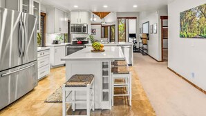 A modern kitchen with white cabinetry, a stainless steel refrigerator, a central island with stools, and a large window providing natural light. - Waikomo Stream Villas 530 – A/C, 2 Bedroom, PBAC Membership (Koloa)