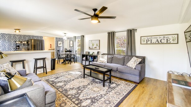A modern living room with a grey sofa, a coffee table, and a patterned rug. There is a kitchen area with a bar and a ceiling fan. - Camp-Style Cabin Near Pigeon Forge: Fire Pit, Deck (Sevierville)