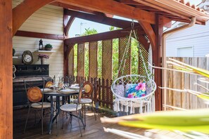 Een houten terras met een hangstoel, een tafel met stoelen en een decoratieve muur met planten en een klok.