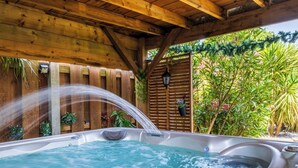 Un bain à remous installé sous une pergola en bois, agrémenté d’un élément décoratif aquatique, et entouré d’une végétation luxuriante.