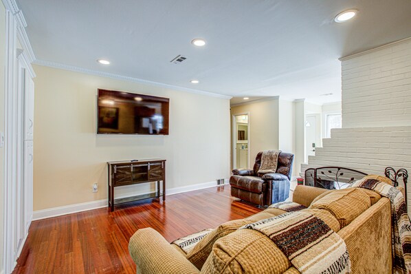 A spacious living room with hardwood flooring, a large flat-screen TV mounted on the wall, a leather armchair, and a sofa with a patterned throw.