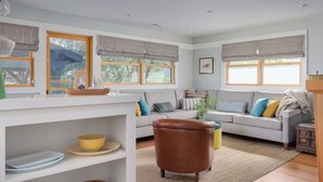 Una sala moderna con sofá, sillón y una mesita de centro. Hay ventanas con cortinas, una estantería y una planta.
