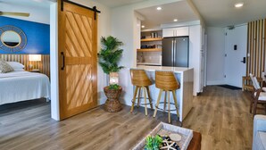 A modern interior with a bed, a dining area with stools, a kitchen with a refrigerator, and a large wooden door. - Completely remodeled 2 bedroom King bed AC Kauhale Makai on the top floor (Kihei)