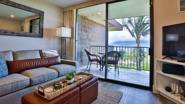 A modern living room with a sofa, coffee table, and a TV. There's a balcony with a view of palm trees and a mountain. - Completely remodeled 2 bedroom King bed AC Kauhale Makai on the top floor (Kihei)