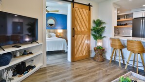 A modern living room with a flat-screen TV, wooden flooring, a sliding barn door, a bed with a blue accent wall, and a kitchen area with bar stools. - Completely remodeled 2 bedroom King bed AC Kauhale Makai on the top floor (Kihei)
