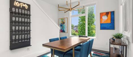 A modern dining area with a wooden table and blue chairs, a wine rack with glasses, and a large window offering a view of greenery.