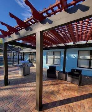 Un área de patio al aire libre con una parrilla, asientos y una piscina al fondo.