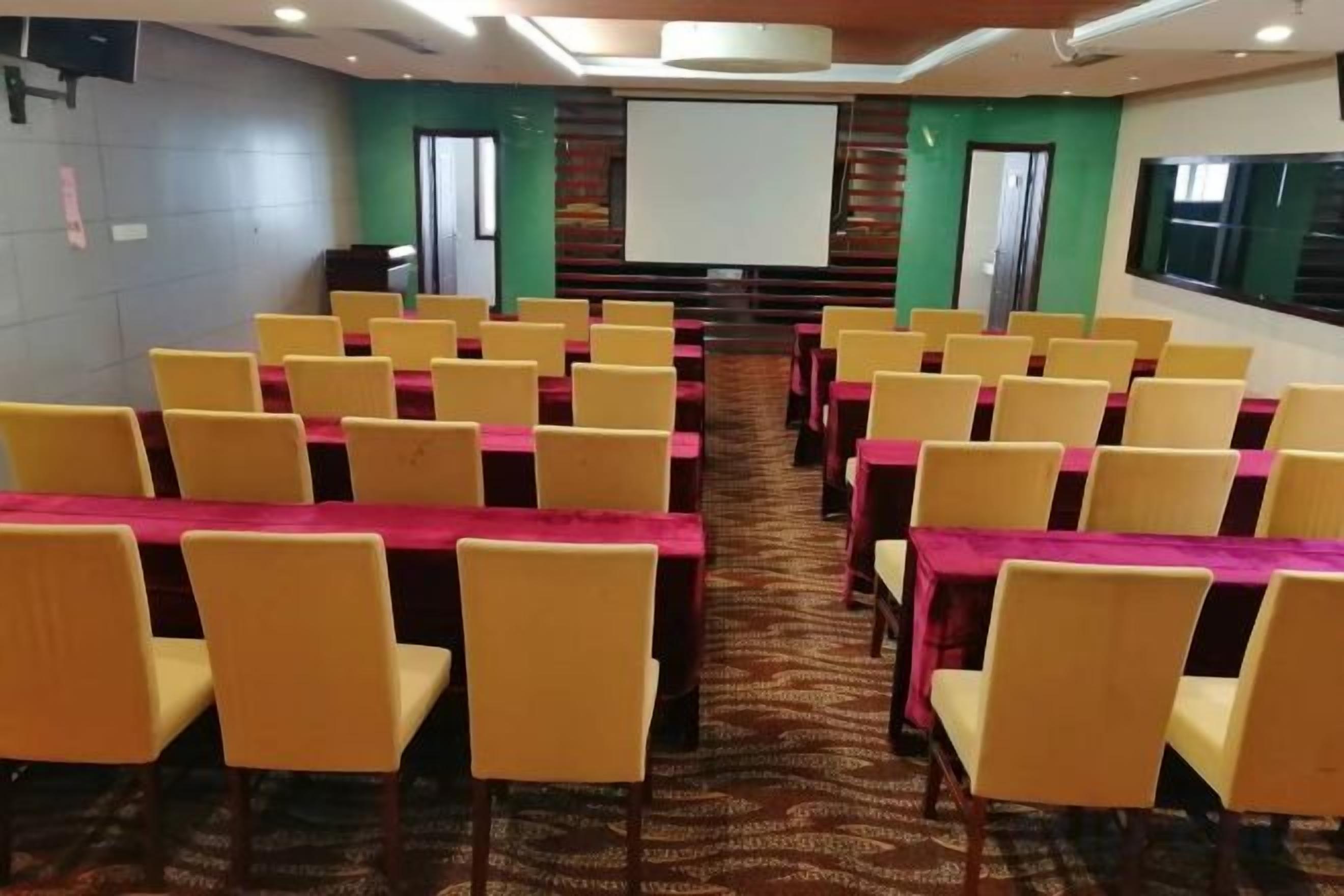 A conference room with rows of yellow chairs and tables, a projector screen, and a carpeted floor.