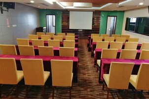 Un salón de conferencias con filas de sillas amarillas y mesas, una pantalla de proyector y suelo alfombrado.