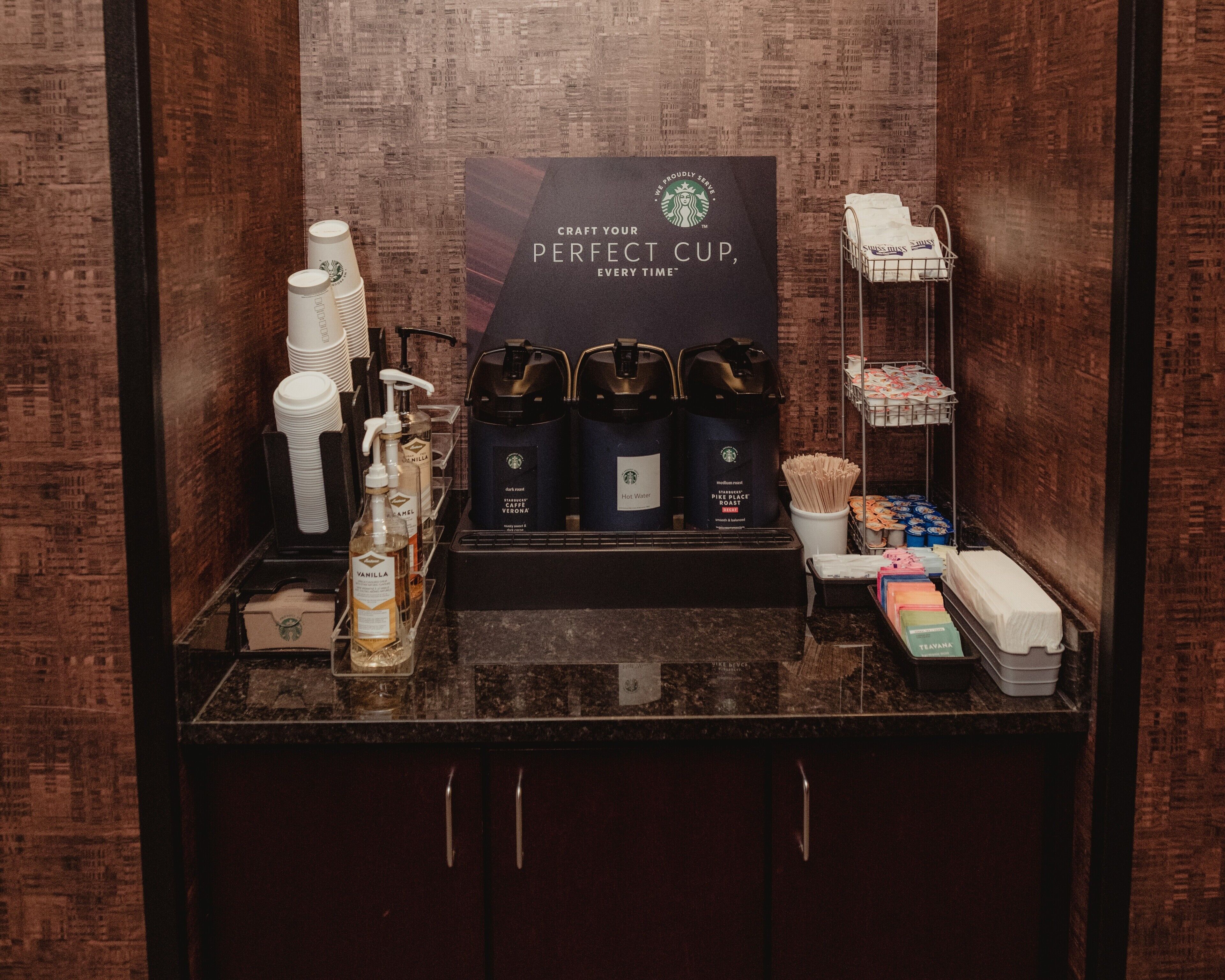 Una estación de café con productos Starbucks, incluyendo tazas, una máquina de café y un cartel que invita a los huéspedes a preparar su taza perfecta.
