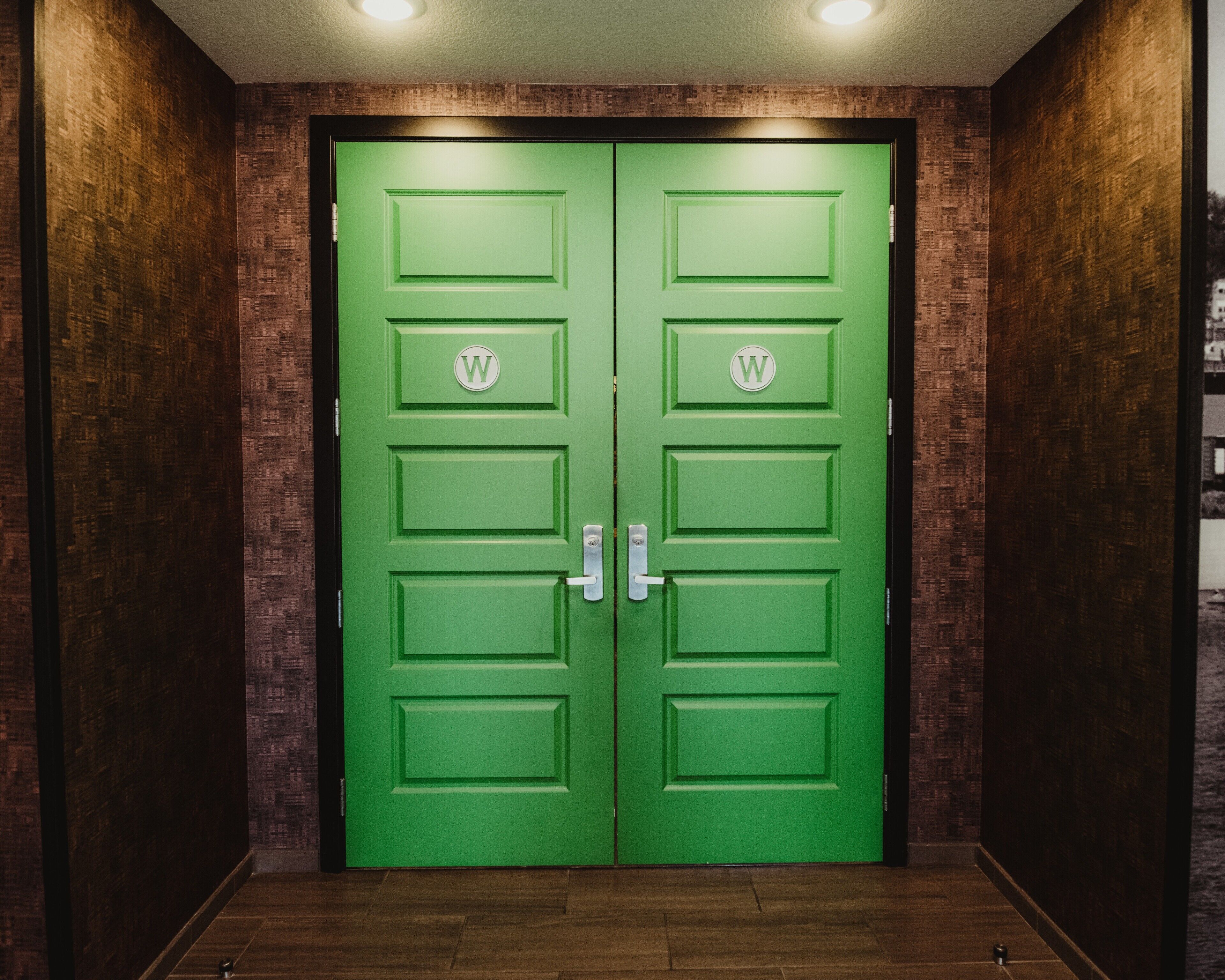 Een groene dubbele deur met een 'W' logo, in een hal met houten wanden en een houten vloer.