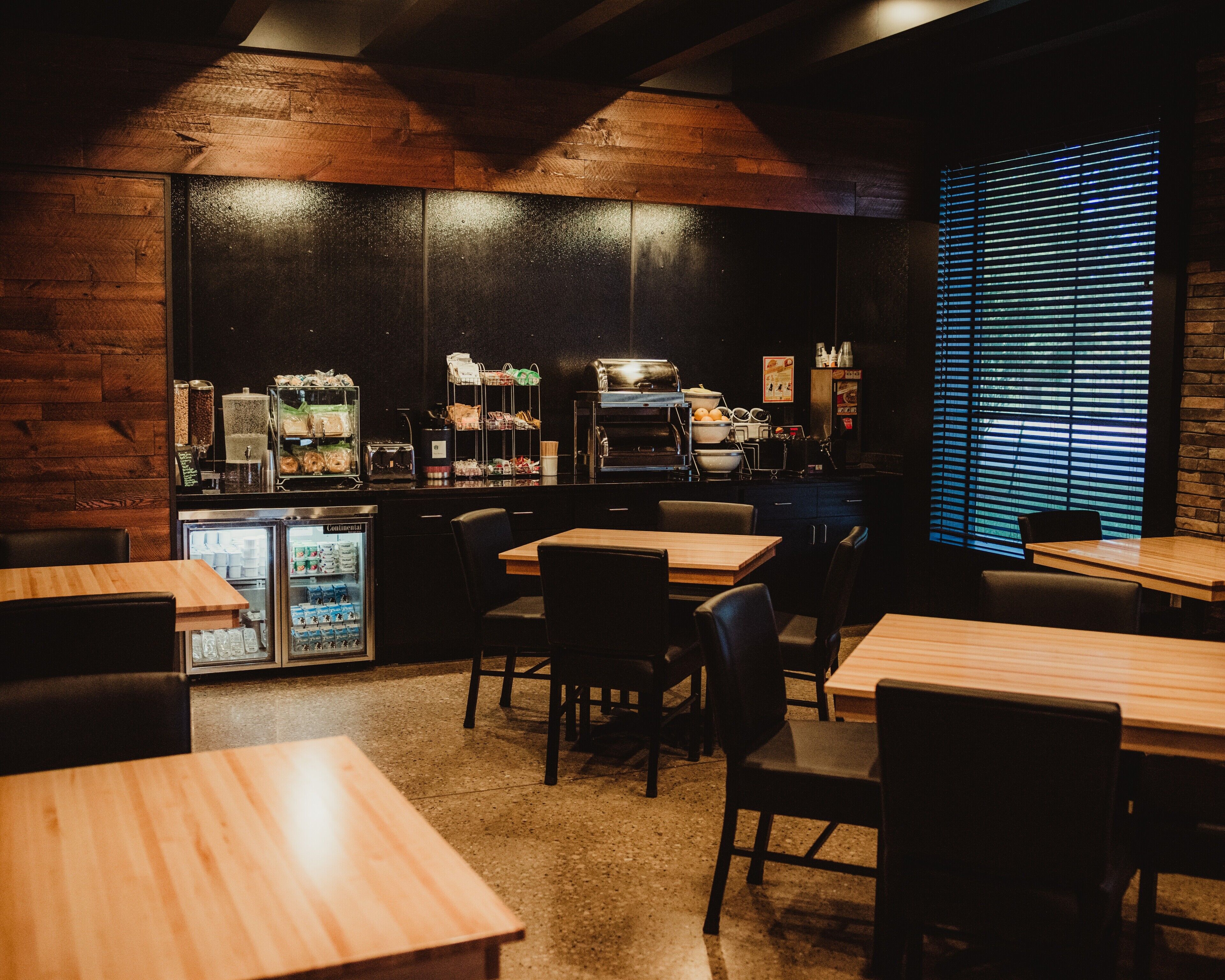 Un restaurante con mesas y sillas de madera, una barra con comida y bebidas, y un refrigerador.