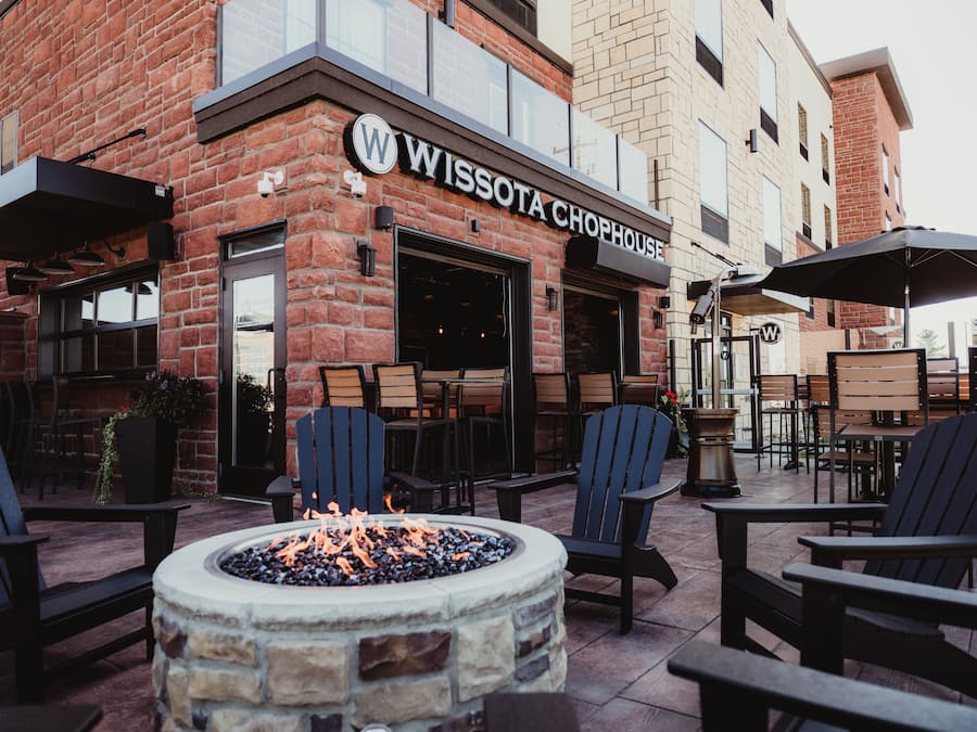 An outdoor seating area with Adirondack chairs and a fire pit, in front of a building with a 'Wissota Chop House' sign.