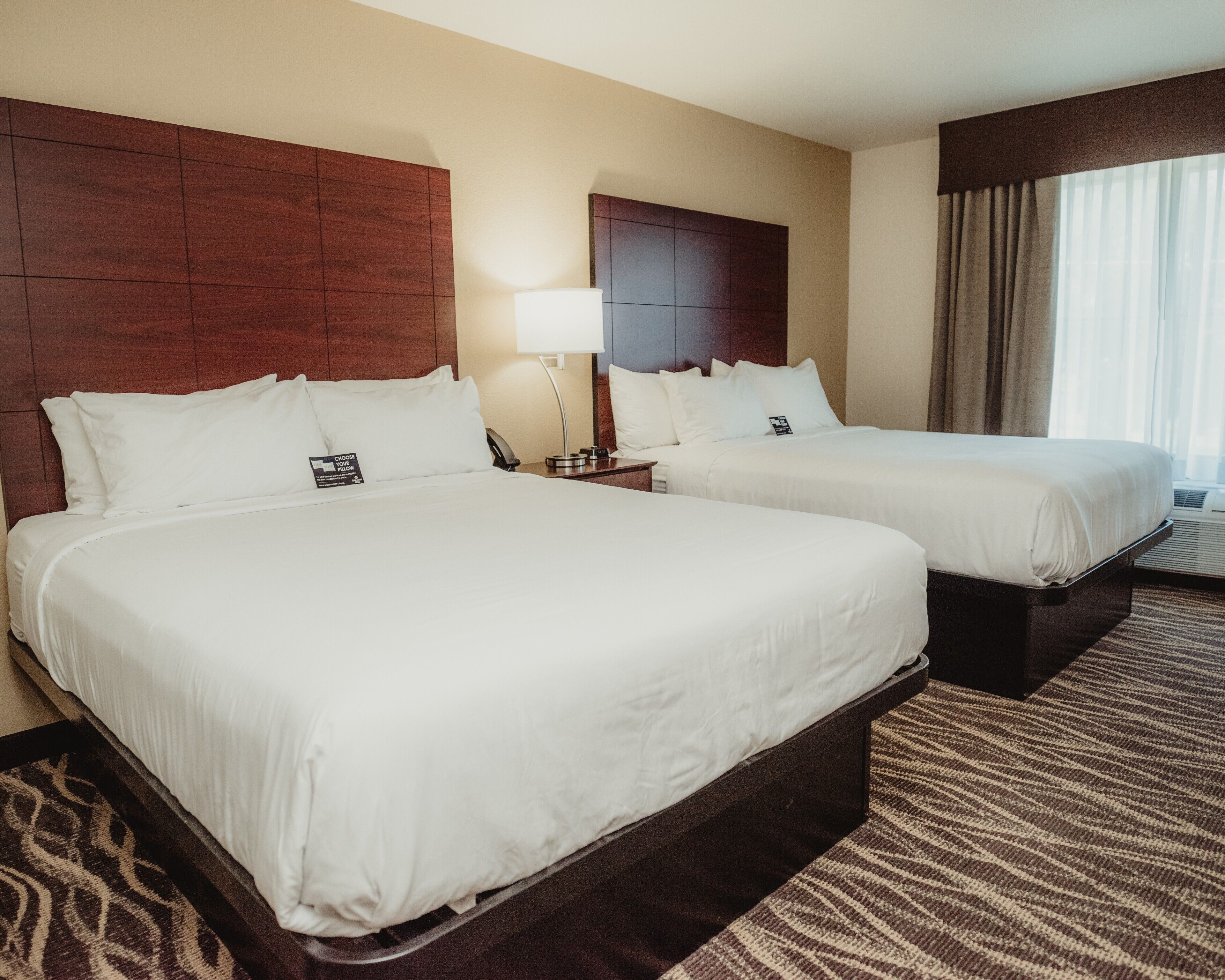 A hotel room with two beds, a nightstand with a lamp, and a window with curtains.