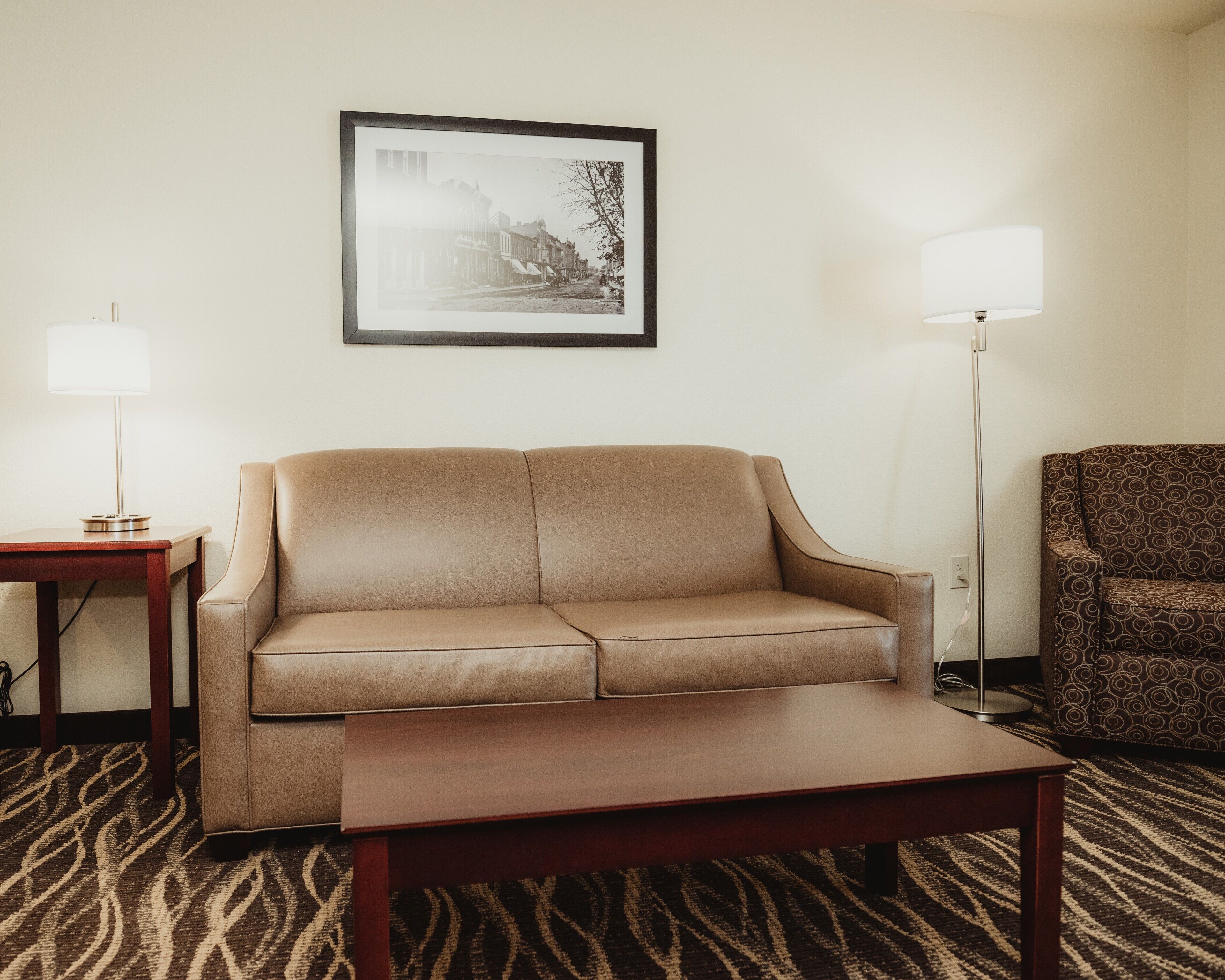 Una habitación de hotel con un sofá color beige, una mesa de centro de madera, un sillón estampado y dos lámparas de pie.