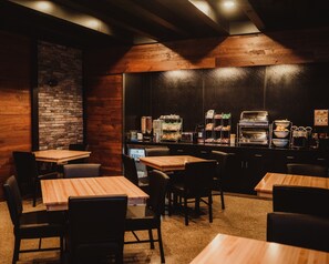 A restaurant interior with wooden tables and chairs, a brick wall, and a counter with a coffee machine and food display.