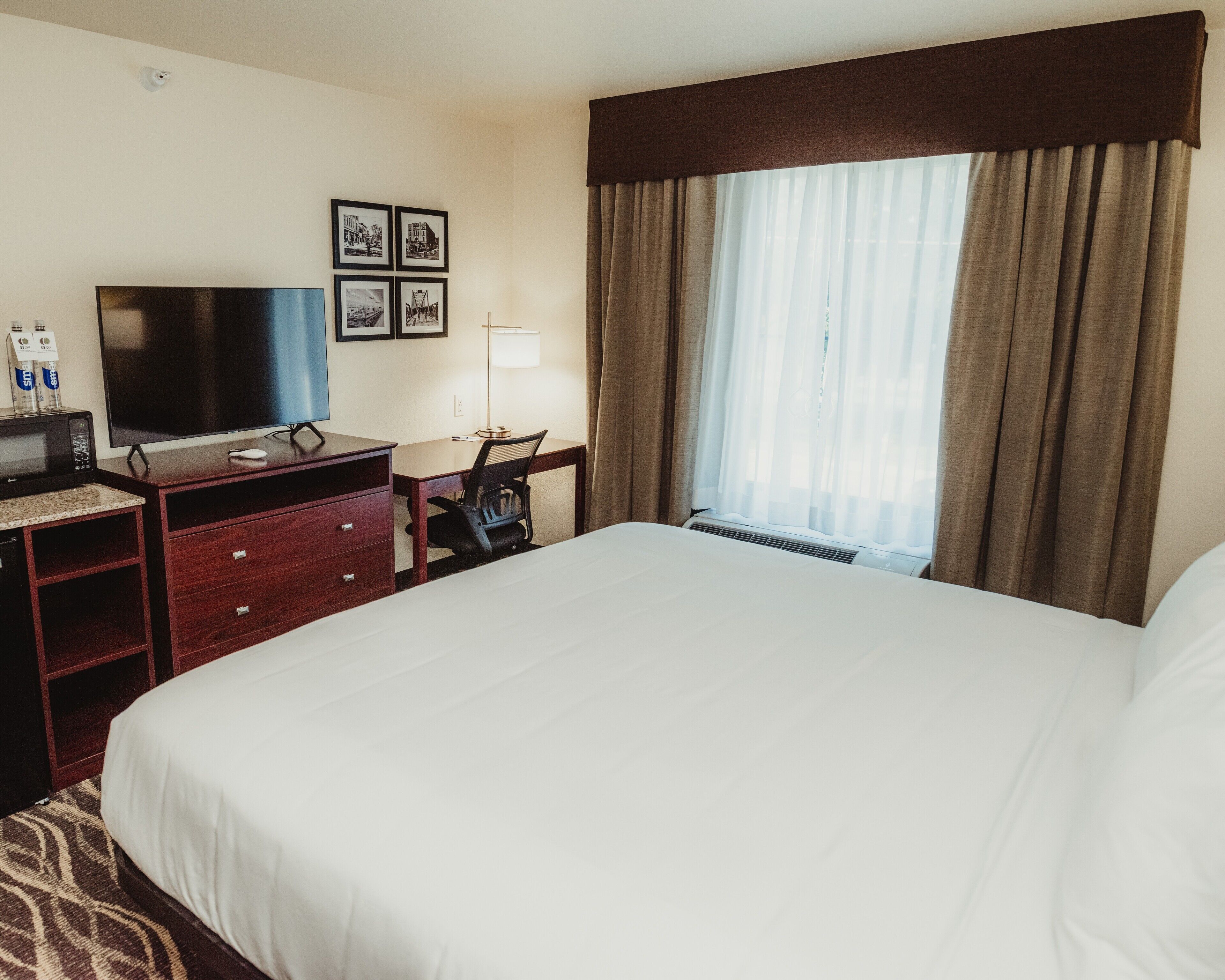 Habitación de hotel con una cama grande, un escritorio con silla, televisor de pantalla plana, lámpara y ventana con cortinas.
