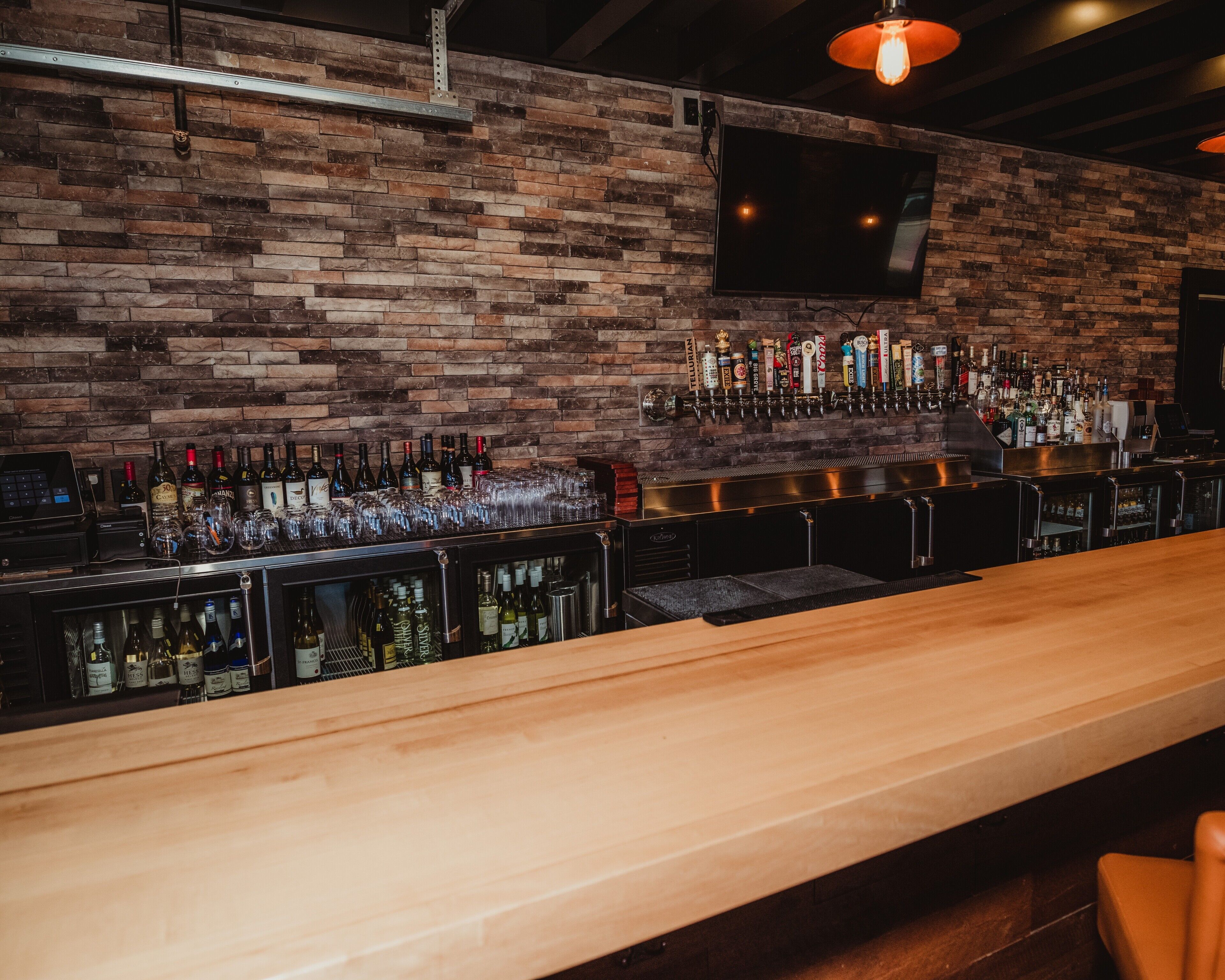 A bar with a brick wall, a flat-screen TV, and a variety of bottles and glasses.