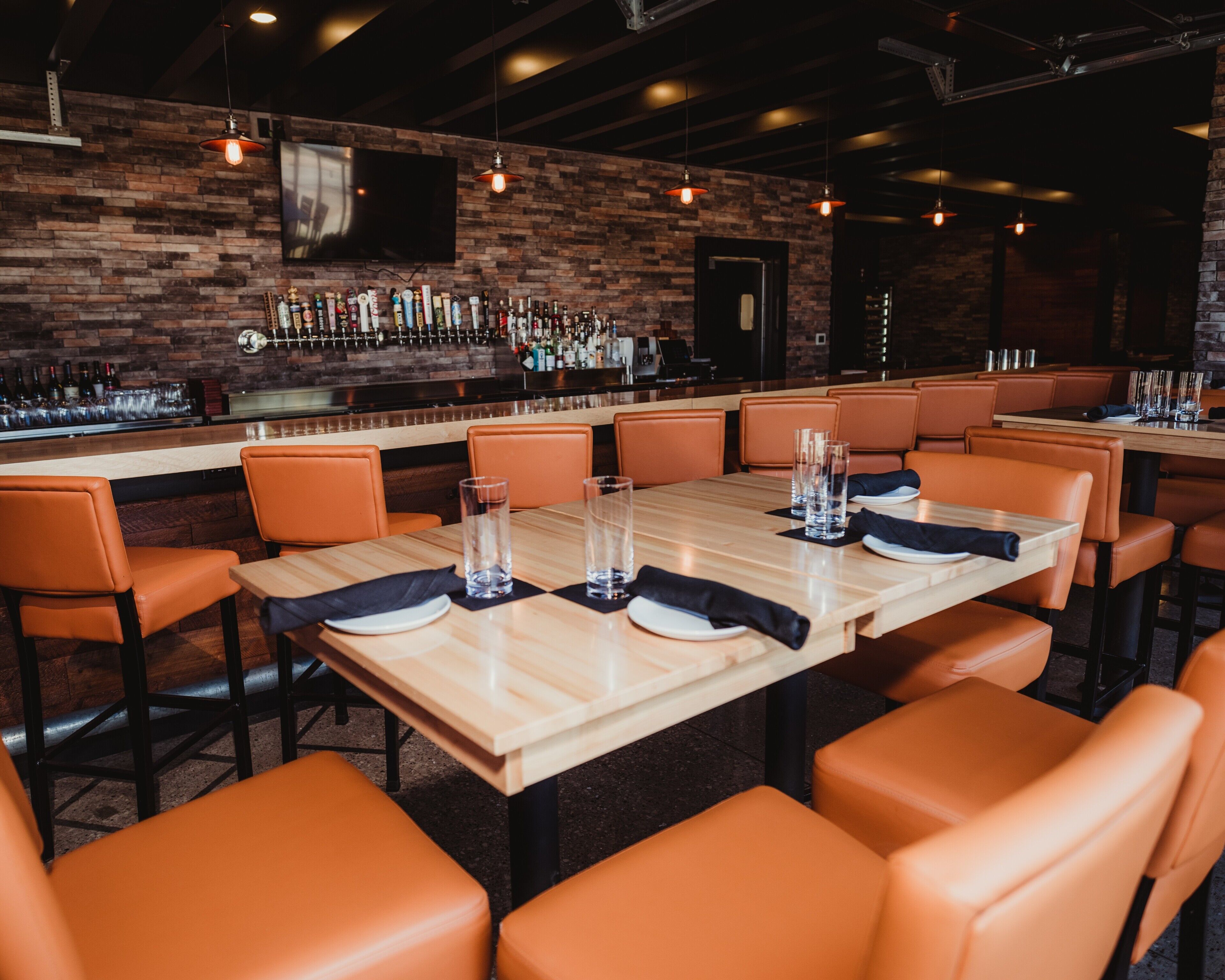 A restaurant with wooden tables and orange leather chairs, a brick wall, and a bar area with bottles.