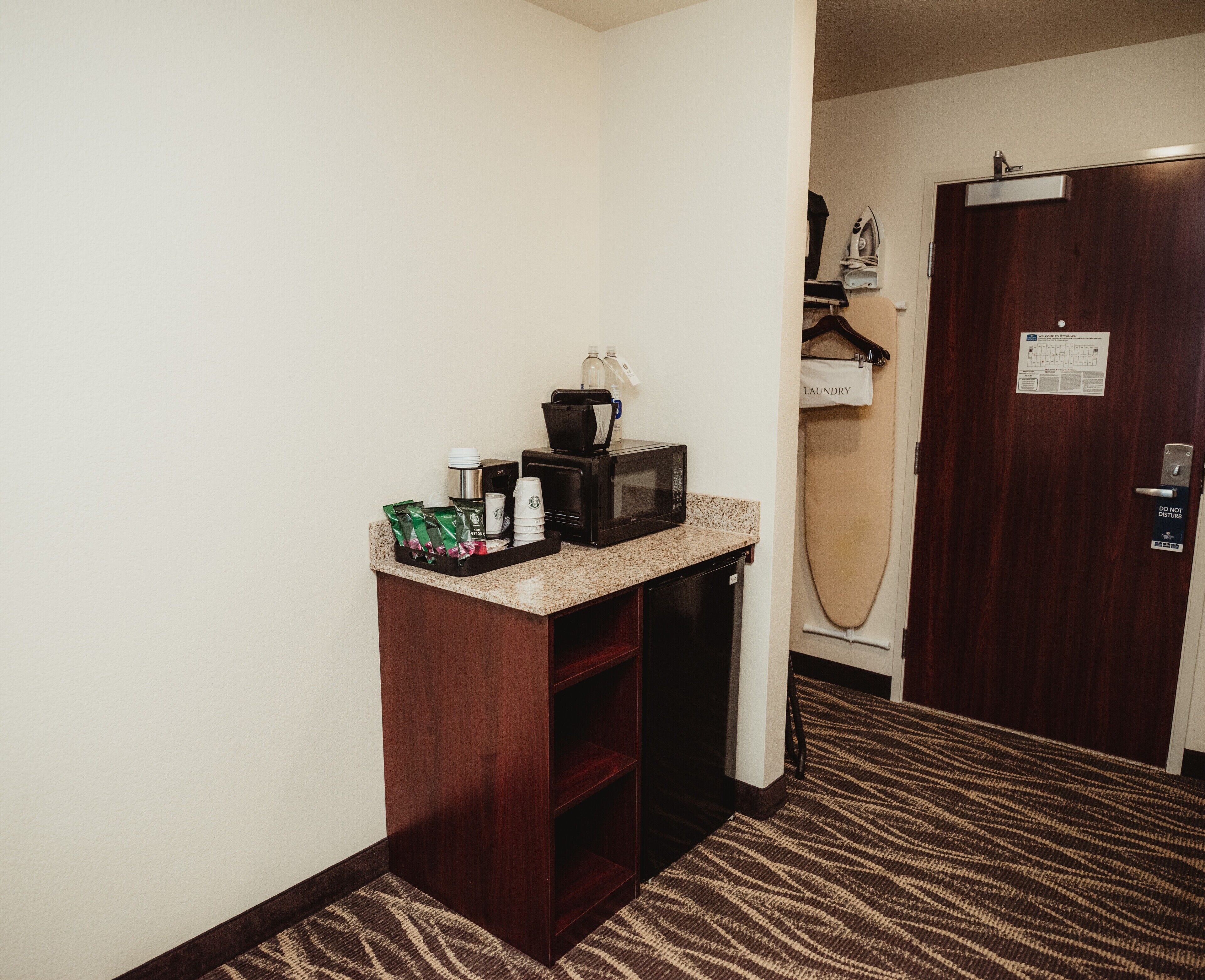 A hotel room with a kitchenette area, including a microwave, coffee maker, and a small refrigerator. There is a door with a 'Do Not Disturb' sign and a laundry sign on the wall.