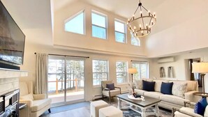 A spacious living room with a fireplace, large windows, and a chandelier. - Designer Beachfront Chalet w/ Theater, Arcade & Beach Firepit Near Schoodic (Gouldsboro)