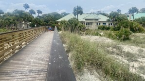 Une promenade en bois mène à un bâtiment surmonté d’un toit végétalisé, entouré de palmiers et de dunes de sable.