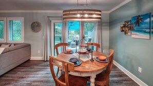 Un coin repas avec une table en bois, des chaises et un lustre. On aperçoit un canapé et une fenêtre avec des rideaux à l'arrière-plan.