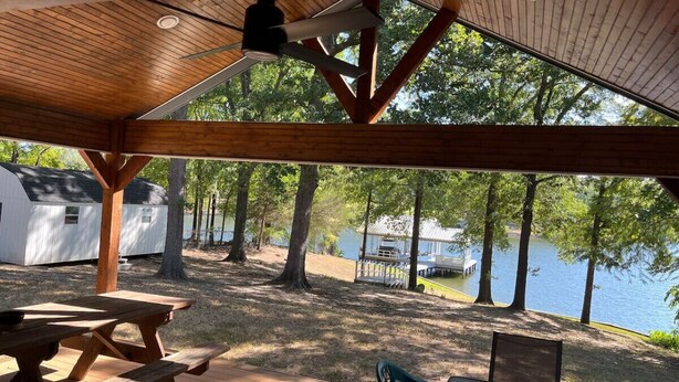 Un espace pique-nique abrité, doté de bancs et de tables en bois, offrant une vue sur un lac avec des chalets en arrière-plan.