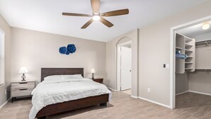 A bedroom with a bed, two bedside tables, a ceiling fan, a wall-mounted light fixture, and a closet with shelves. - Your Home away from Home (Goodyear)
