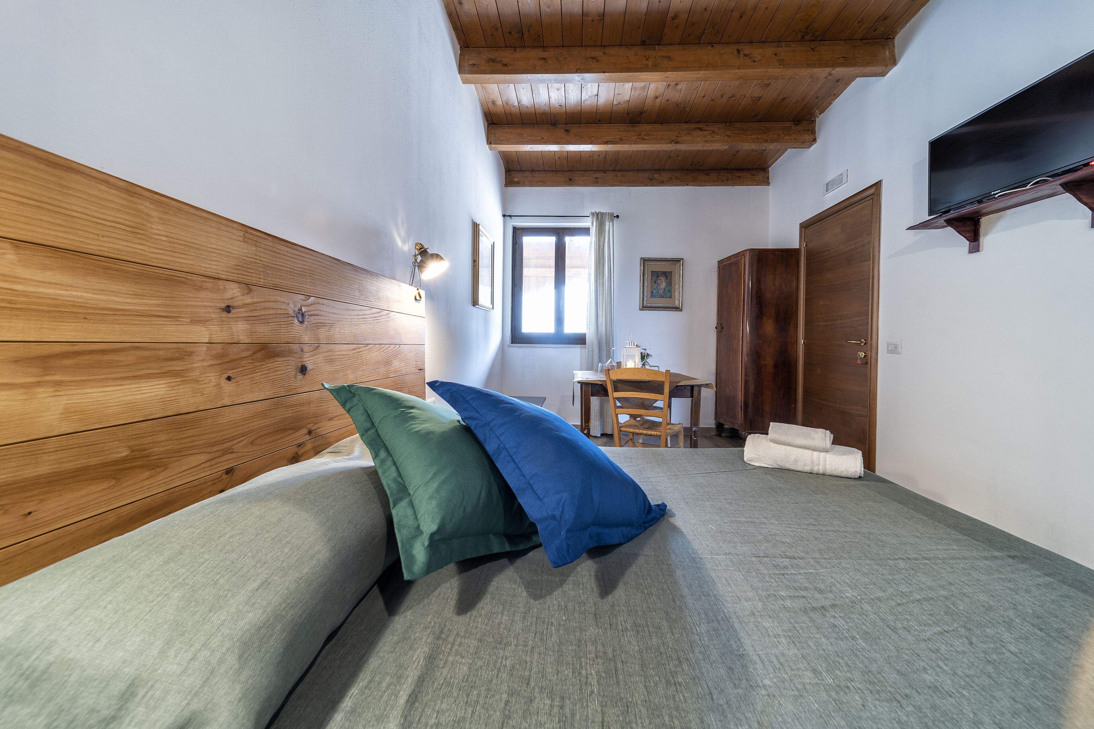 Quarto com cabeceira de madeira, cama com almofadas verdes e azuis, televisão de tela plana na parede e janela com cortinas.