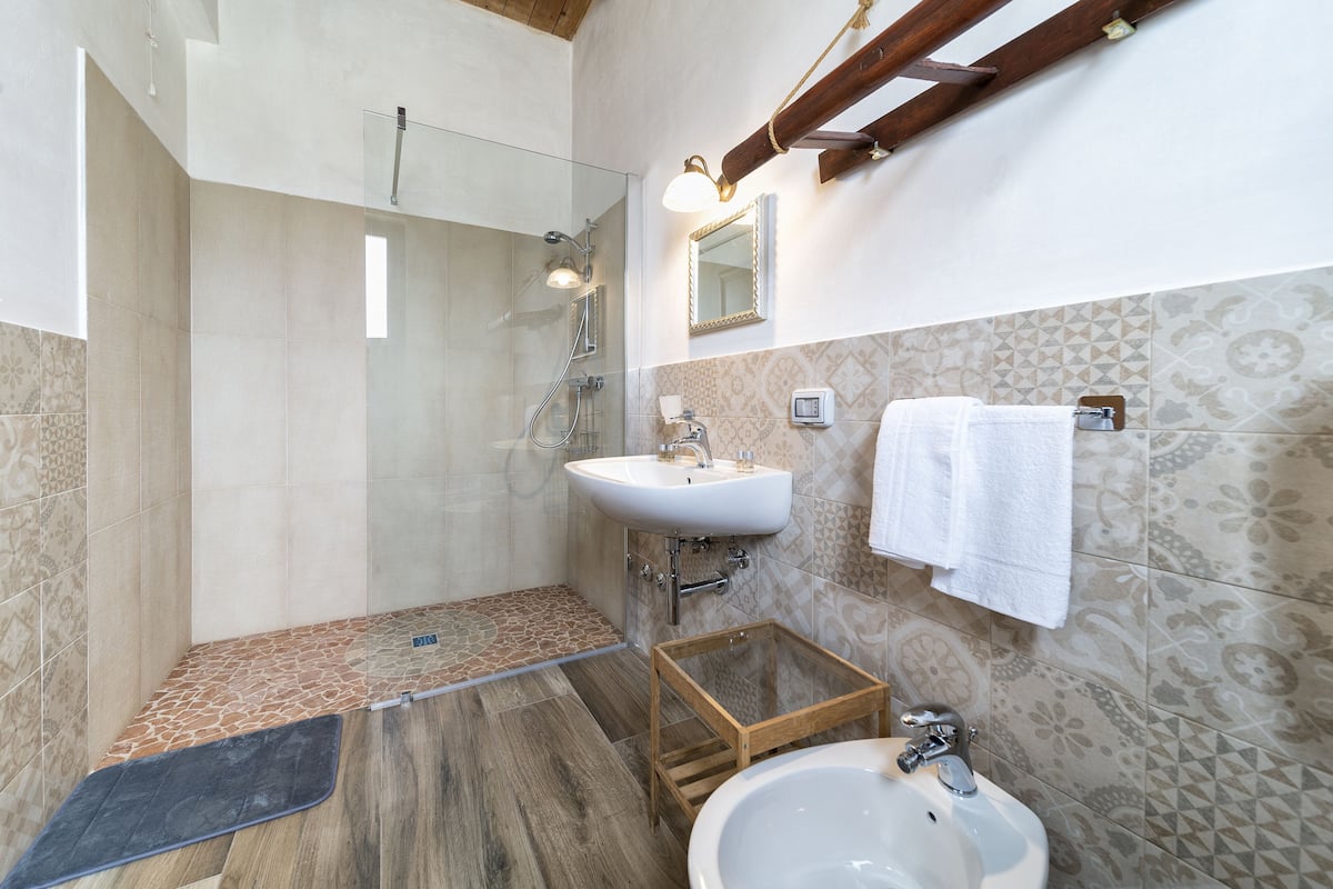 Un baño con cabina de ducha de vidrio, lavamanos, espejo y un taburete de madera.