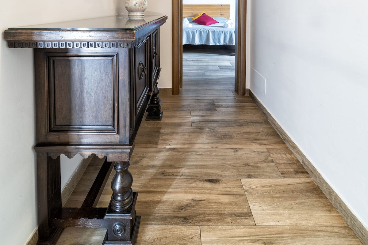 Un pasillo con piso de madera, un aparador de madera oscura, un cuadro enmarcado y una cama a lo lejos.