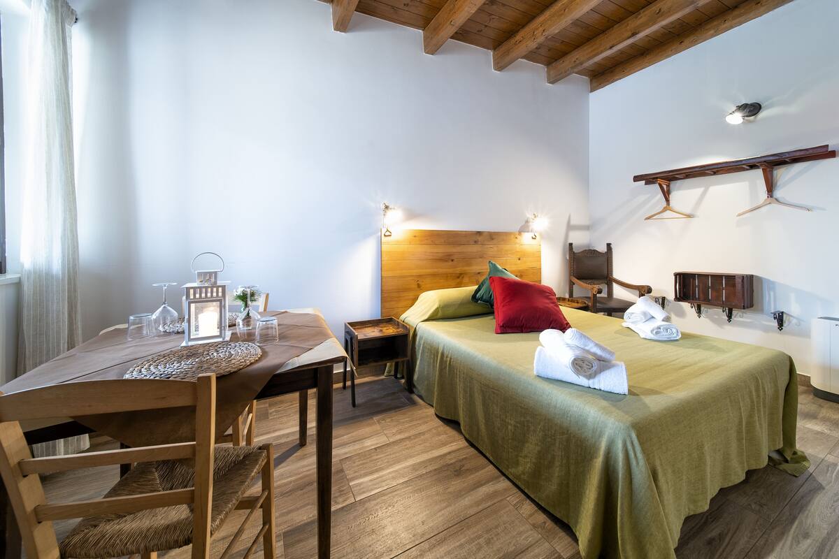 Una habitación con una mesa de madera, una cama con cubrecama verde, cabecero de madera y un perchero en la pared.