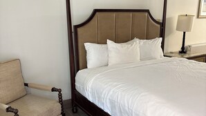 A four-poster bed with white linens, a wooden chair with a beige cushion, and a nightstand with a lamp. - Richland Inn (Johnstown)