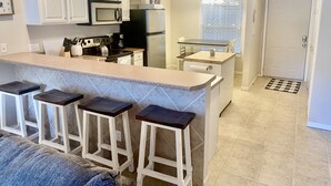 Une cuisine moderne avec des meubles blancs, un îlot central doté de tabourets de bar, un réfrigérateur et un coin repas avec une table et des chaises en verre.