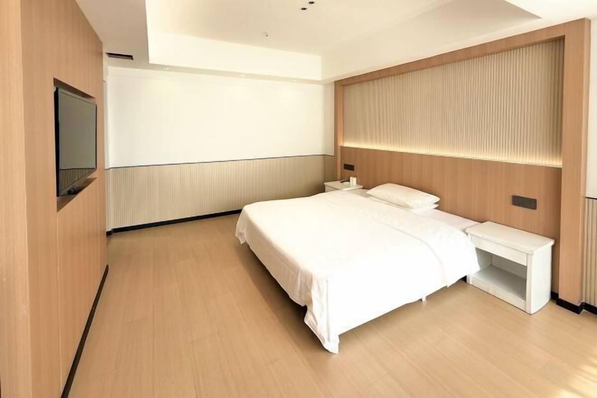 A modern hotel room with a large bed, a bedside table, a TV mounted on the wall, and a wooden headboard.