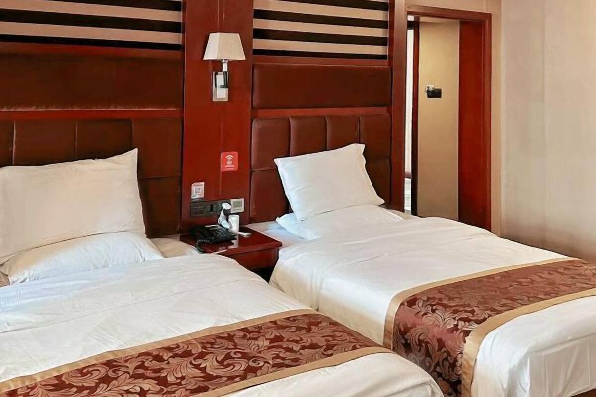 A hotel room with two beds, a wooden headboard, a bedside table with a lamp, and a red emergency exit sign.
